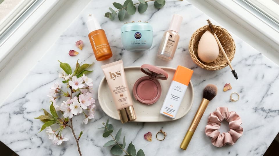 Flat lay of spring beauty products including tinted moisturizer, cream blush, and vitamin C serum on marble background
