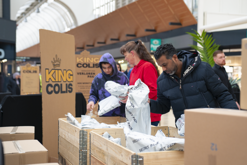 People opening Secret Pack parcels from a wooden crate at a King Colis lost packages event in a shopping centre.
