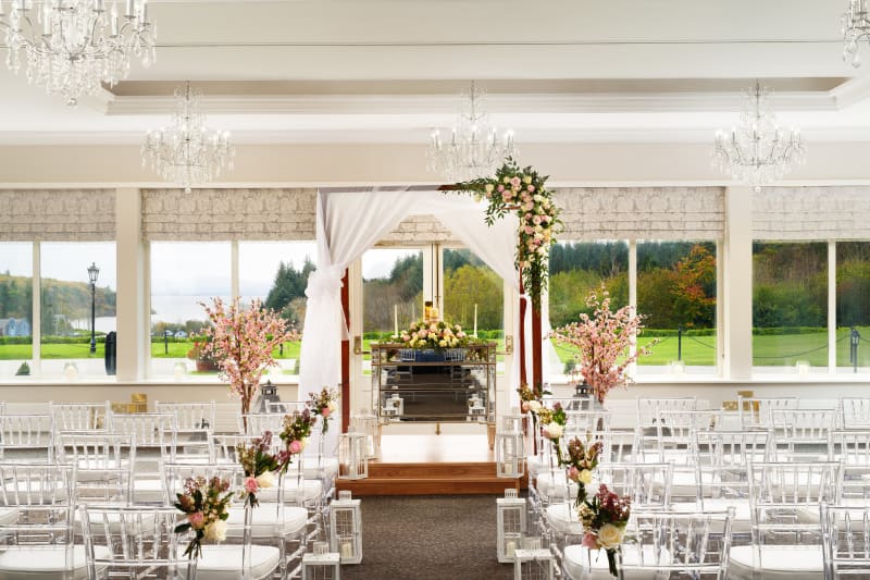 Elegant indoor wedding ceremony room with floral arch, ghost chairs, chandeliers and garden views.