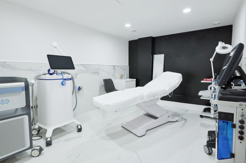 Modern aesthetic clinic treatment room with white treatment bed, laser device, and multiple skin treatment machines.
