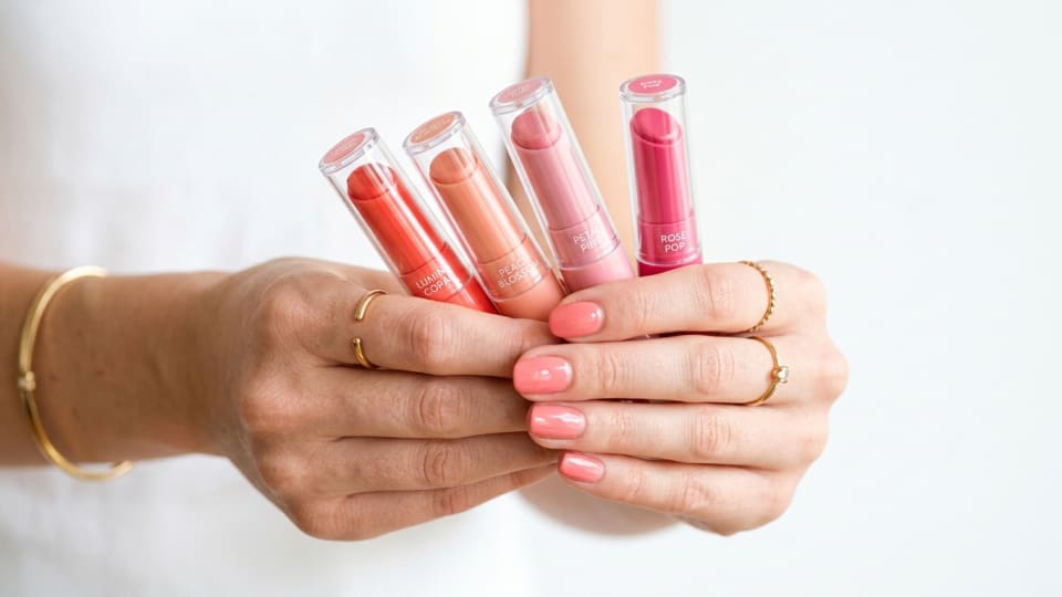 Close-up of hands holding spring lip colors in coral, peach, and pink shades against a bright white background