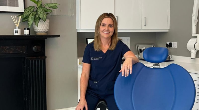 Dr Jennifer Collins in navy dental scrubs beside a blue dental chair at Northumberland Dental Care.