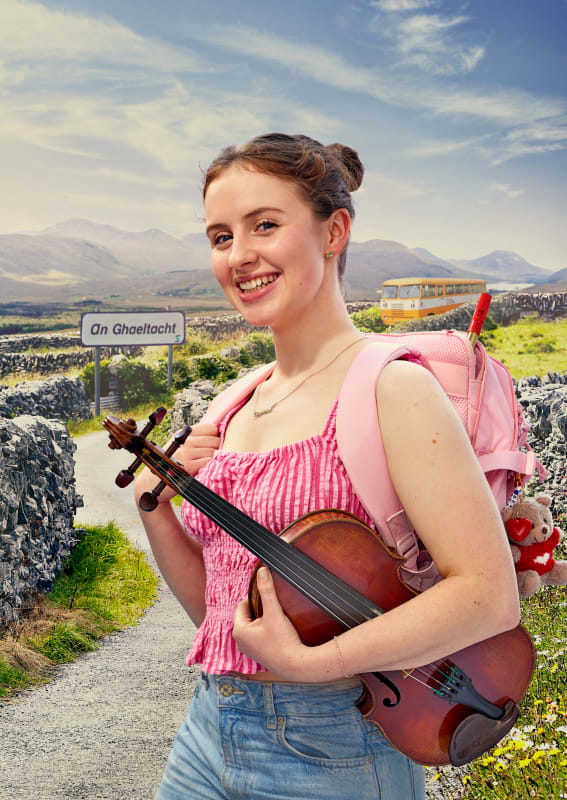 Saoirse smiling outdoors at An Ghaeltacht sign, holding a violin, wearing pink top and carrying a backpack.