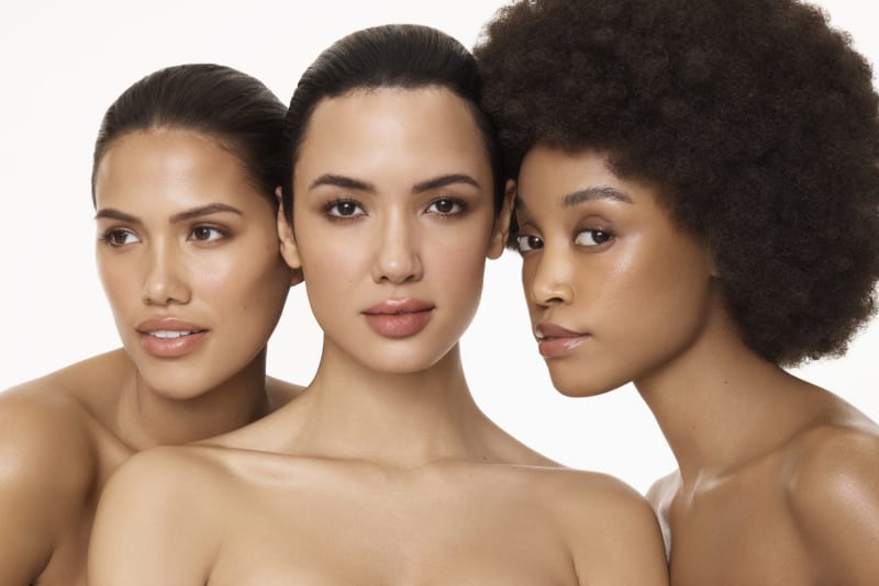 Three women with diverse skin tones and glowing complexions posed together in a beauty campaign shot.