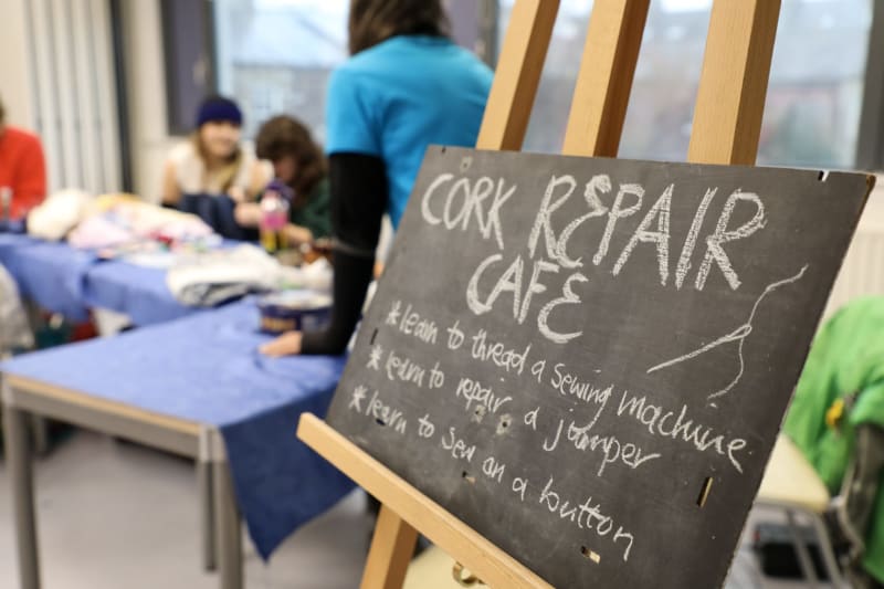 Cork Repair Cafe chalkboard sign listing sewing skills, with people working on fabric repairs in background.
