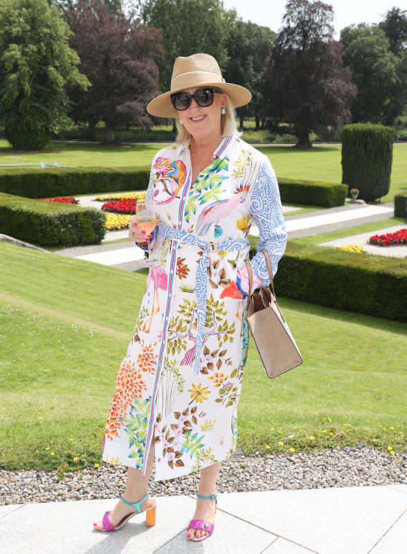 Paul Costelloe wearing a tropical-print midi shirtdress, straw hat and colourful sandals in a formal garden.