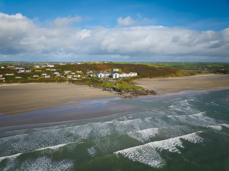 Aerial view of a sandy Irish beach with rolling waves, a small coastal village on a headland, and green fields beyond.