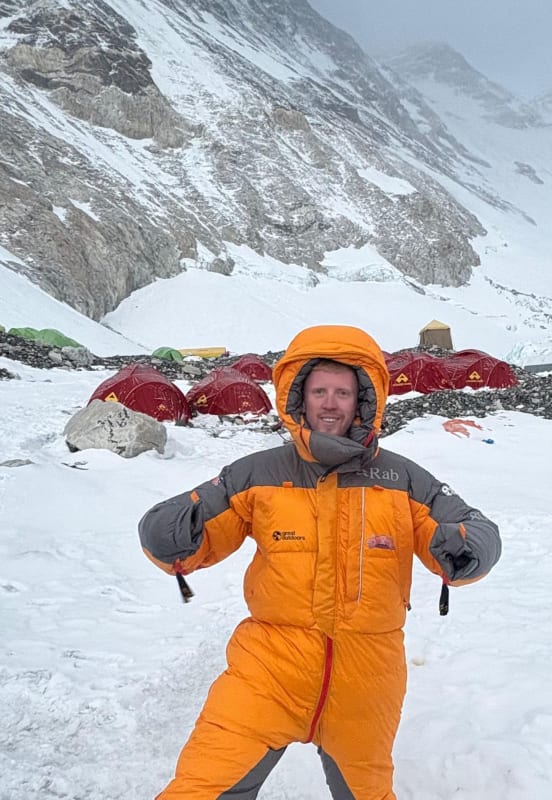 Adam Sweeney in an orange Rab down suit at a high-altitude mountain base camp surrounded by red tents.