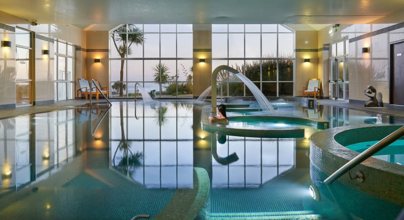 Indoor spa pool with circular hydrotherapy jets, large coastal windows, and a person relaxing at dusk.
