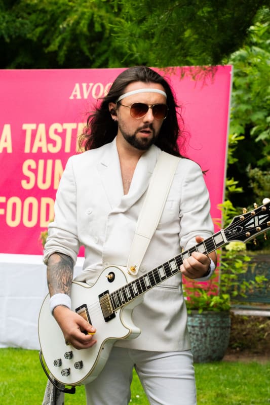 Musician in white suit playing a white Les Paul guitar at an Avoca summer food tasting event outdoors.