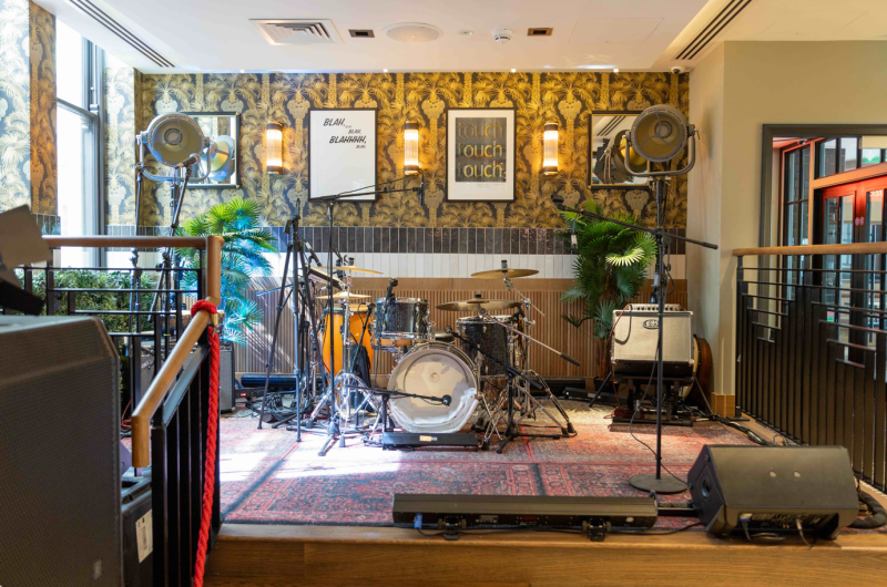 Small live music stage with drum kit, mic stands, and monitor speakers in a stylish bar with botanical wallpaper.