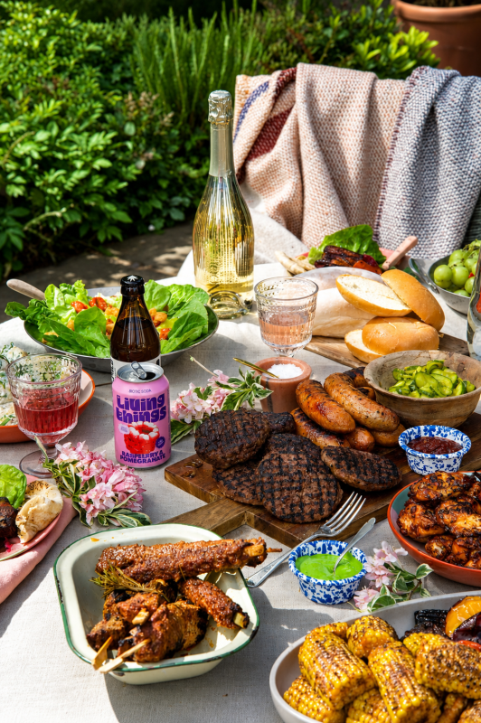 Outdoor garden table loaded with barbecue food including burgers, sausages, wings, corn, salad, rolls, and drinks.