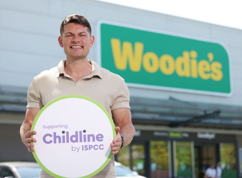 Man holding a 'Supporting Childline by ISPCC' sign outside a Woodies store.