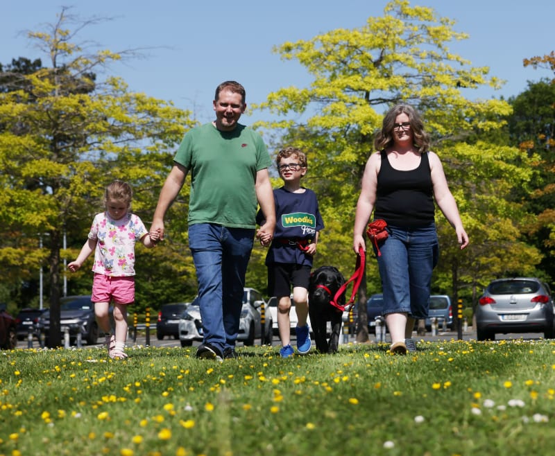 Family with two children and a black Labrador assistance dog walking on grass on a sunny day.