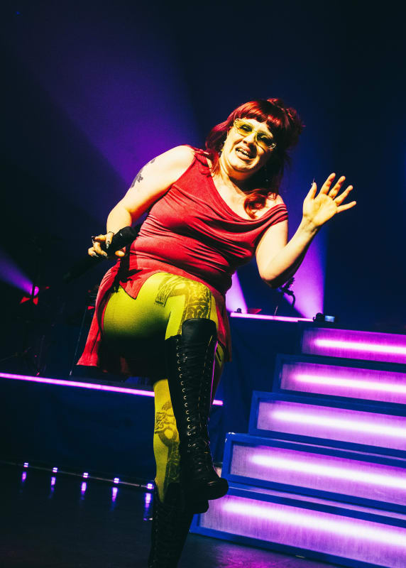 Lainey performing on stage in a red top, yellow-green leggings and black thigh-high boots under purple stage lighting.