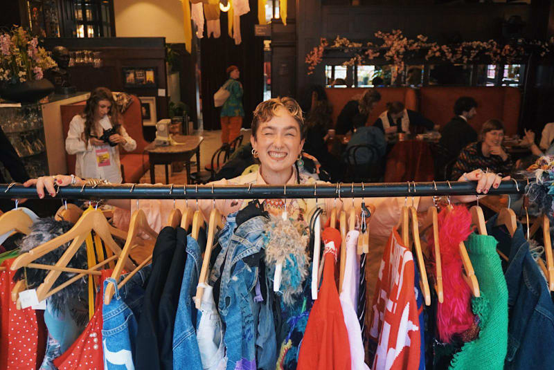 Nicole Holbrook smiling behind a rail of colourful vintage clothing at an indoor market event.