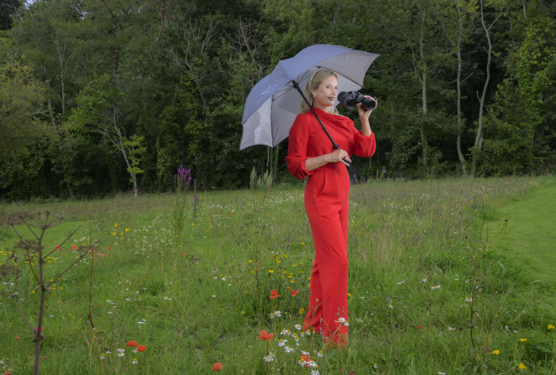 Sarah Butler stands in a wildflower meadow in a red jumpsuit, holding a silver The Grace umbrella and binoculars.
