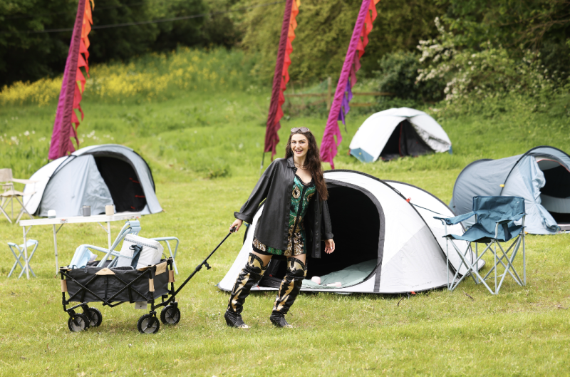 Woman celebrating at a festival campsite beside a dome tent and a 'No Tent Left Behind' campaign sign.