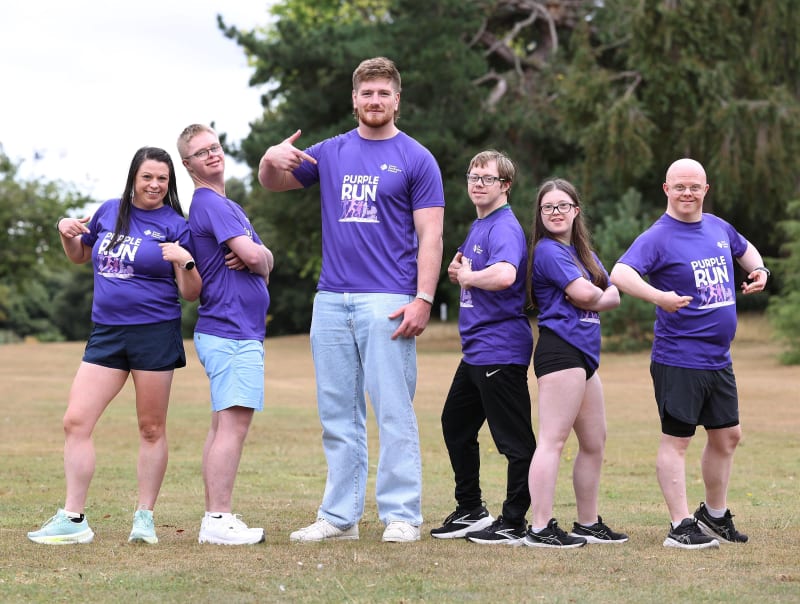 Six people in purple 'Purple Run' t-shirts posing outdoors on grass, four of whom have Down syndrome.