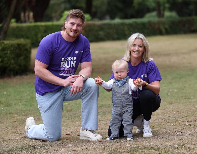 Two adults in purple Down Syndrome Ireland Purple Run t-shirts help a baby stand between them in a park.