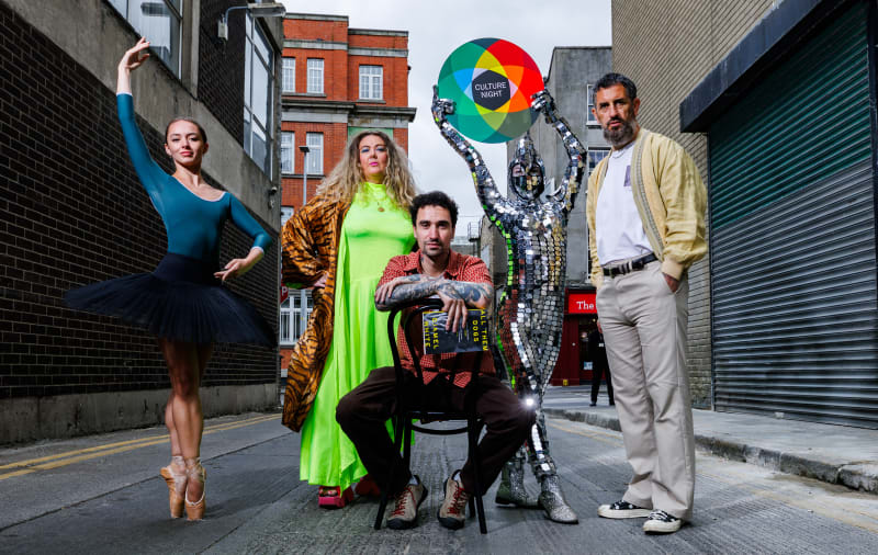 Five performers pose in an urban alleyway to promote Culture Night, including a ballet dancer and a mirror-costumed figure.