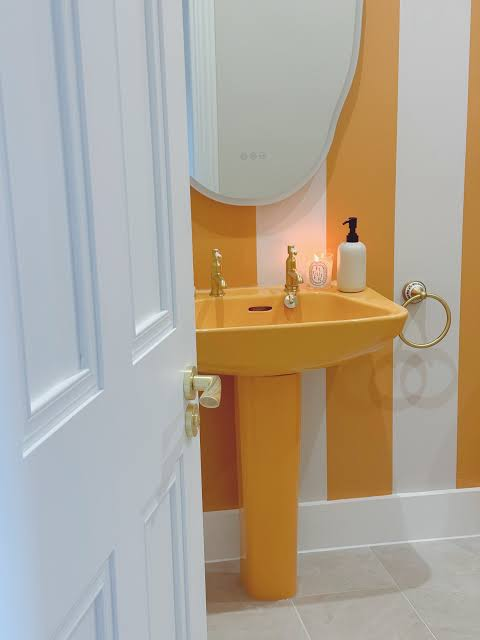Yellow pedestal sink with brass taps in a bold orange and white striped powder room with oval mirror.