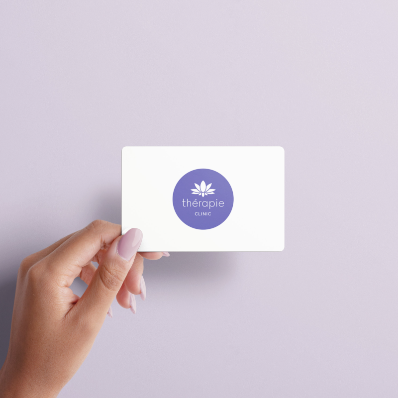 Hand holding a white Thérapie Clinic gift card with purple lotus logo against a lavender background.