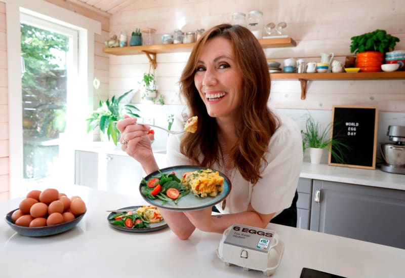 Woman smiling in a styled kitchen eating a frittata slice, with free range eggs and a World Egg Day sign visible.